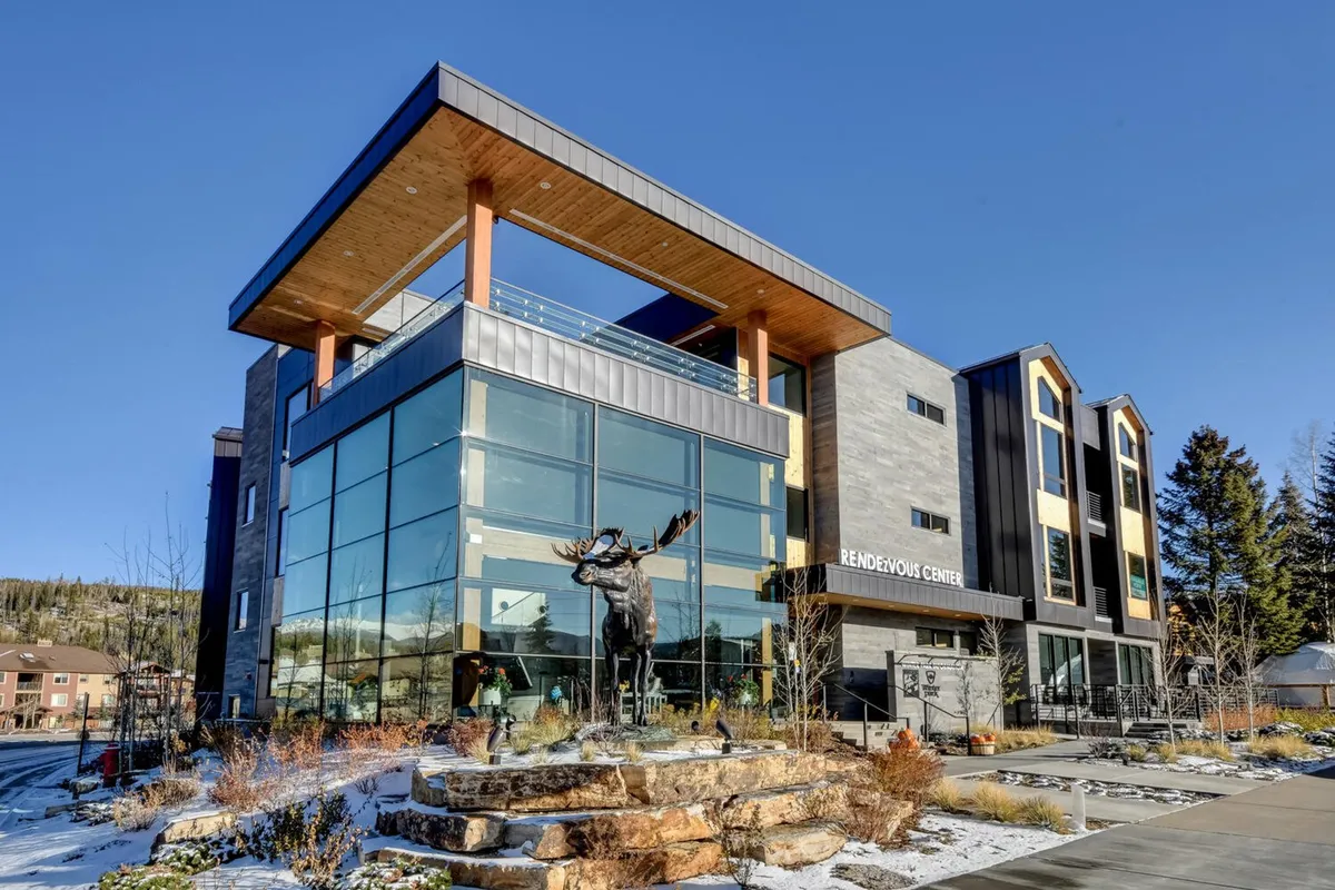 A stylish exterior shot of a modern building with a large glass facade, a whimsical moose statue, a wooden overhang, and thoughtfully landscaped gardens. The scene exudes a sense of community and open space, ideal for walks and outdoor activities.