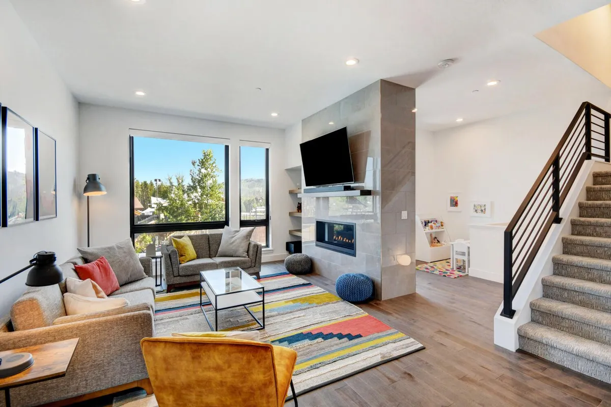 A comfortable seating area arranged around a sleek, modern fireplace, with large windows framing sweeping views of the outdoors. The colorful area rug and inviting furnishings create a welcoming space, perfect for family gatherings or entertaining guests. The ambience is cozy yet contemporary, and the expansive windows make the scenic mountain or park views a focal point.