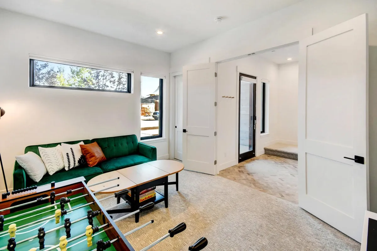 A sunny, playful game room distinguished by a green sofa, a foosball table set up near large windows that allow natural light to flood the space, and an open doorway suggesting flow into other areas. People are engaged in friendly foosball matches and cozy conversations on the sofa, suggesting both activity and relaxation happen here.