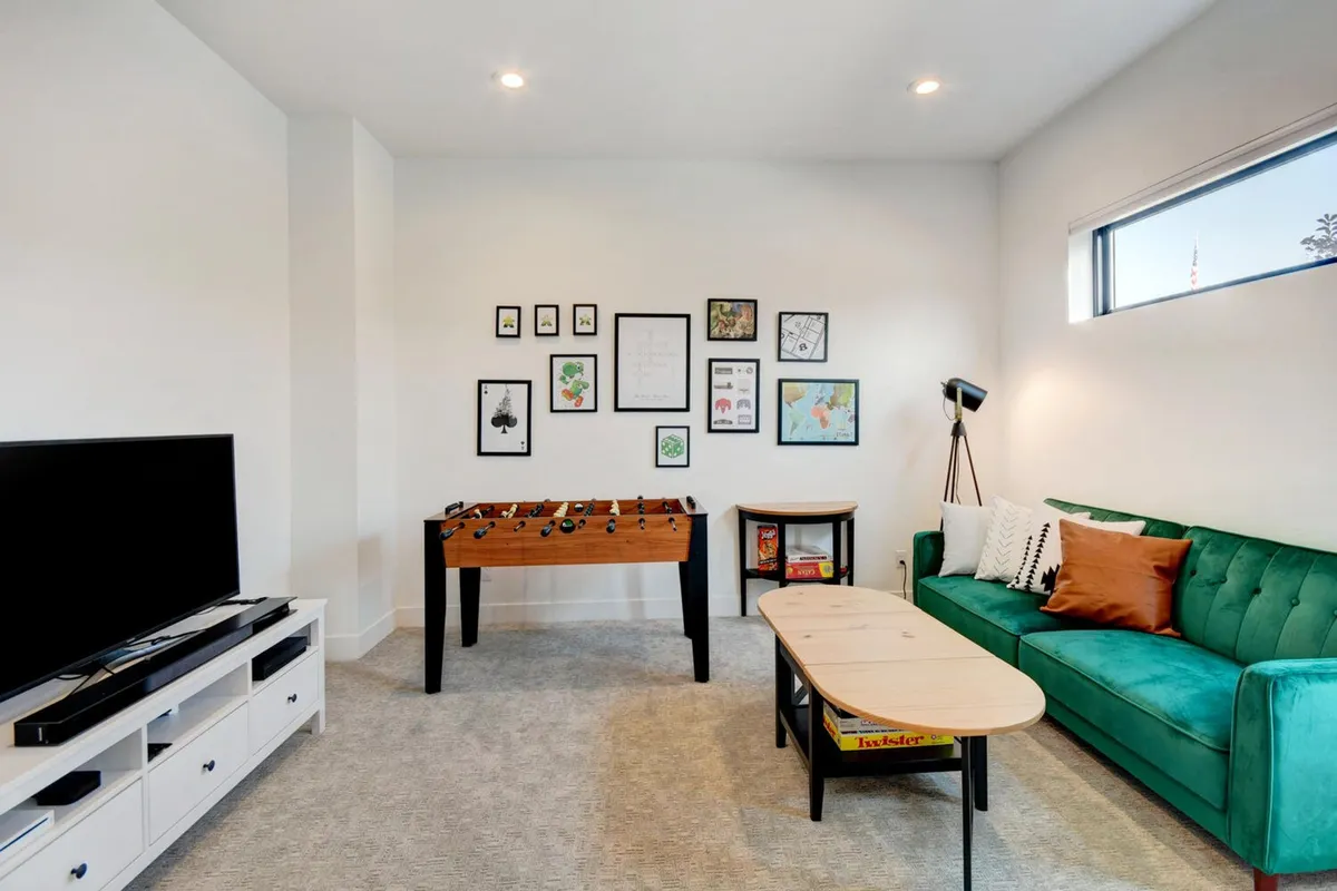 A vibrant game room featuring a green couch, a foosball table as the centerpiece, a stylish gallery wall with framed art, and a functional coffee table. The space is alive with playful activities: a foosball match in progress, someone relaxing on the couch, and areas that invite reading or drawing. The overall atmosphere is fun and relaxed, suggesting a versatile room ideal for both active play and winding down.