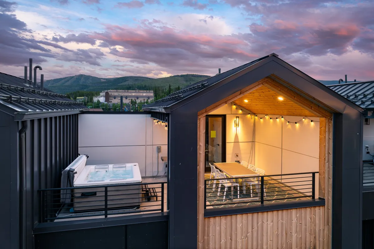 A cozy rooftop deck featuring a bubbling hot tub, an outdoor dining table set for an evening meal, glowing string lights overhead, and panoramic mountain views in the background. The warm ambiance is perfect for relaxing, stargazing, and intimate gatherings under the open sky.