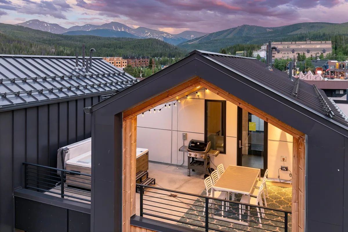 A stylish rooftop deck with a hot tub, string lights, and an outdoor dining area, all set against a stunning mountain backdrop. The relaxed atmosphere is perfect for unwinding after a day of adventure, featuring spaces for socializing, dining, and stargazing.