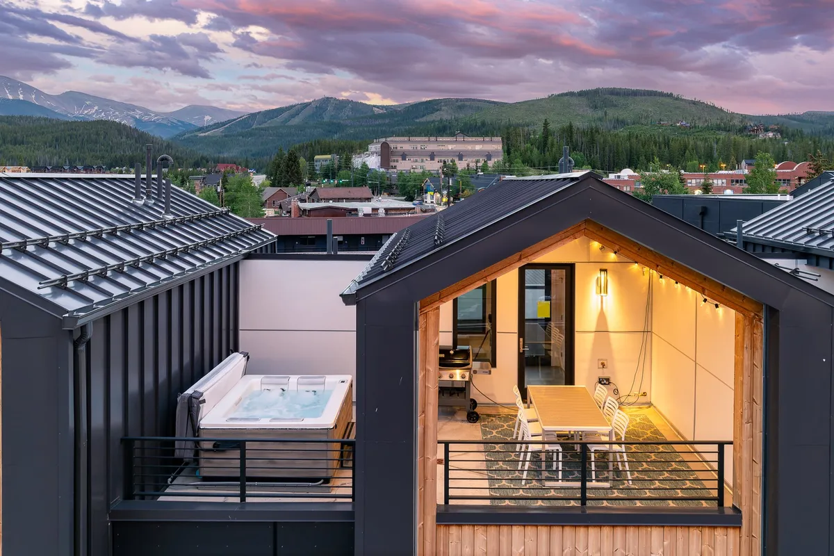 A rooftop deck with a hot tub and outdoor dining area overlooks sweeping mountain views. The tranquil, luxurious setting hints at private enjoyment of public events and sunsets, suggesting the convenience of viewing concerts or festivals from home while highlighting the upscale amenities available to residents.