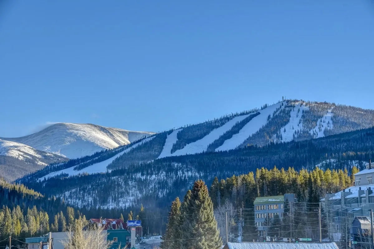 A sweeping view of snow-covered mountain slopes dotted with skiers and snowboarders, pine forests, and the charming town at the base. The image conveys both the majesty of the Rockies and the proximity of world-class skiing to the accommodations.