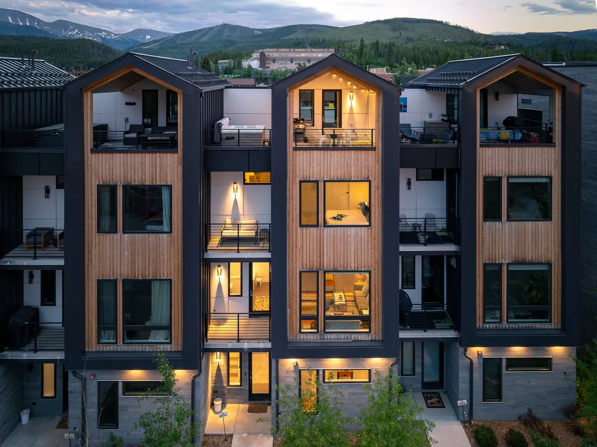 The image showcases the exterior of a new luxury single-family home in Winter Park, Colorado, with three distinctive gabled roofs, expansive windows aglow with inviting warm light, and spacious balconies—some featuring guests enjoying the sunset and mountain vistas. The scene captures a cozy yet grand atmosphere, blending mountain modern architecture with the comfort of illuminated interiors and the promise of vibrant social spaces outside.