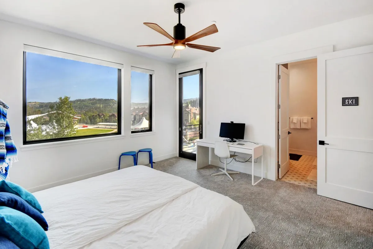 A cozy bedroom featuring large windows that frame beautiful views, a comfortable bed with layered blankets, and a desk with a computer. The room is bathed in natural light, offering both relaxation and a practical area for work or reading.