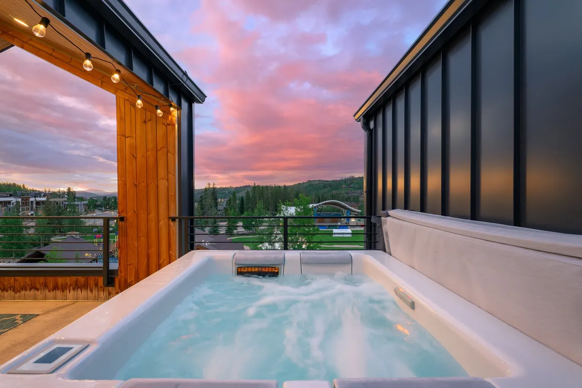 A modern hot tub perched on a rooftop deck, facing a vast mountain landscape and a vibrant sunset sky. The scene is tranquil, inviting viewers to imagine unwinding after a day of adventure while taking in the breathtaking surroundings.
