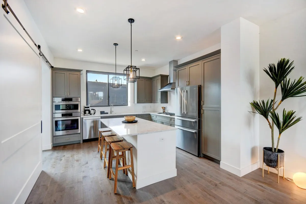 A spacious, light-filled kitchen featuring a large central island, stylish pendant lighting, and top-of-the-line modern appliances. The setup invites socializing and culinary creativity, with ample room for guests to gather and enjoy casual dining or conversation. The indoor plant adds a touch of natural warmth to the space.