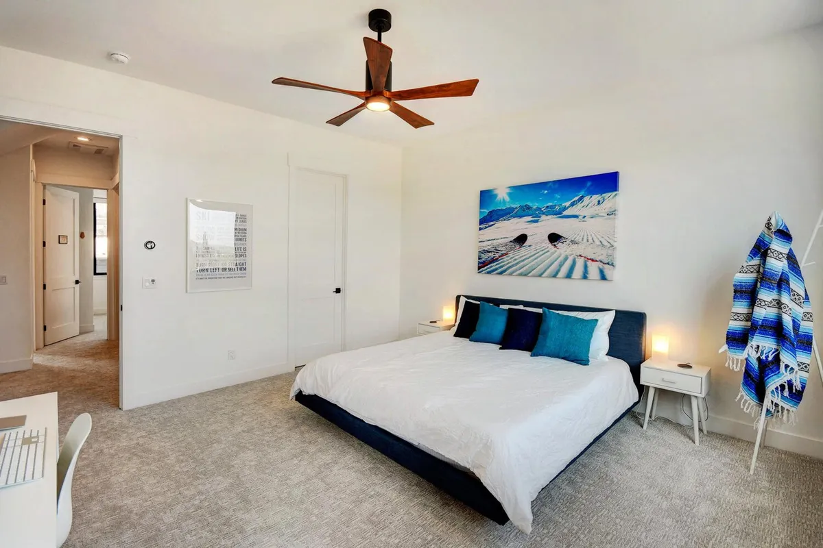 A tranquil bedroom with a plush bed dressed in blue and white bedding, accompanied by mountain-themed artwork, a modern ceiling fan, a decorative throw chair, and small bedside tables with lamps. Soft natural light filters through the window, creating a peaceful retreat. The presence of a desk hints at a functional workspace within the serene setting.