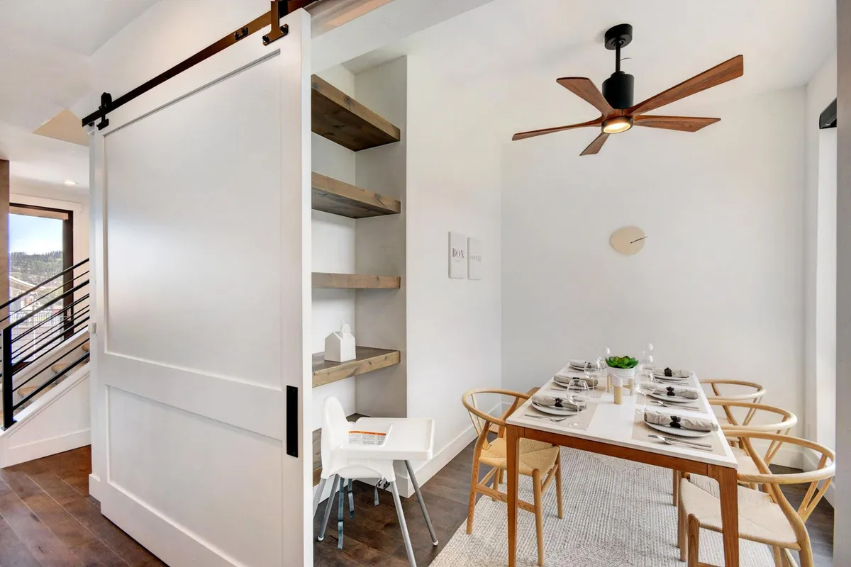 An elegant dining table is set and ready for a meal, surrounded by cozy chairs and warm wooden accents. The space is styled for both intimate family dinners and lively gatherings, with a sliding barn door and open shelving lending a blend of rustic and contemporary charm. Natural light fills the space, suggesting a seamless connection to the living and kitchen areas.