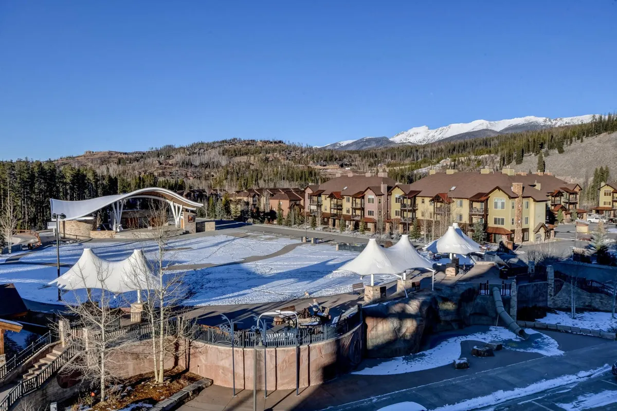 A vibrant exterior scene featuring an open-air ice rink, colorful buildings, mountain backdrop, and festive white tents. The area is bustling with energy, suggesting family outings, winter festivals, and community events that appeal to all ages.